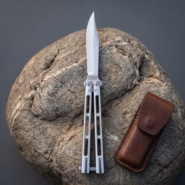 Skeletonised steel butterfly knife with a clip point blade and knotwork etching open on a smooth grey river boulder beside a brown leather pouch