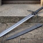 Skull sword with a blackened cast skull guard and chevron etched blade lying on worn cobbles beside a matching black leather scabbard with two belt straps