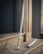 Slender hand forged brass hilted sabre with leaf shaped quillons and a pale leather wrapped grip standing on granite steps beside a cream leather scabbard