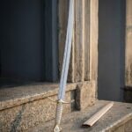 Slender hand forged brass hilted sabre with leaf shaped quillons and a pale leather wrapped grip standing on granite steps beside a cream leather scabbard