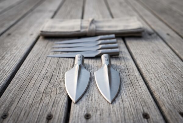 Socketed spearheads stacked on grey decking, two broad leaf heads in front and four narrow heads behind a canvas roll