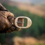 Solid brass belt buckle with an engraved vegvisir on a brown leather belt worn at the waist, gloved hand at the side over a hillside