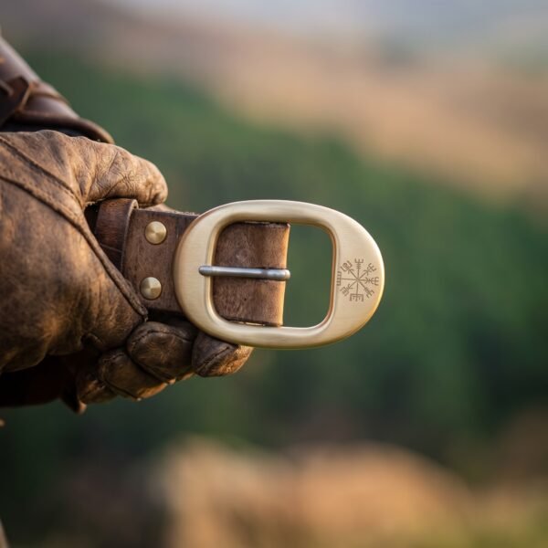 Solid brass belt buckle with an engraved vegvisir on a brown leather belt worn at the waist, gloved hand at the side over a hillside