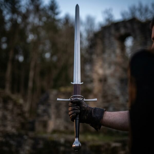 Spring steel montante greatsword held point up in a gloved fist, showing the ricasso and long quillons against forest ruins