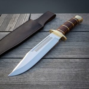 Stacked leather bowie knife with a mirror polished 1095 clip point blade, brass double guard and knotwork etching lying on grey barn boards beside a brown sheath