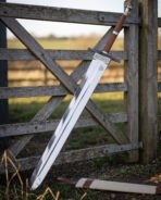 Stacked leather grip arming sword in 1095 steel angled against a farm gate showing the cut knotwork band above the blackened crossguard and turned pommel