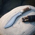 Stag handle kukri knife in polished 1095 steel resting on a smooth grey boulder beside its brown leather sheath holding two small stag handled companion blades.