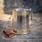 Stainless steel and brass tankard with Celtic knotwork band and riveted body standing on flagstones beside a brown leather belt hanger