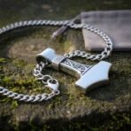 Stainless steel Mjolnir pendant with a knotwork bail and heavy curb chain coiled on mossy stone beside a grey pouch
