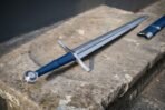 Steel arming sword with blue leather grip, straight cross guard and rune cut fuller resting on a worn stone step