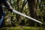Steel arming sword with blue leather grip held by a gloved hand in a mossy woodland, blade pointing to the ground