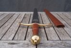 Close up of a steel arming sword from its brass ball pommel end, red leather grip and brass trefoil guard, on a grey wooden table with a tan scabbard