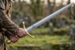 Steel arming sword with a brass trefoil guard and longship etched blade held by a man in a rough tunic in front of a ruined stone wall