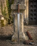 Steel arming sword with engraved arched crossguard and tan leather grip leaning on a carved stone pedestal, scabbard and gloves at its foot