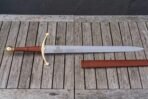 Steel arming sword laid flat on grey wooden slats with a longship etched on the blade, brass ball pommel, red leather grip and tan leather scabbard