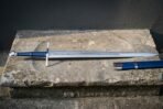 Steel arming sword laid flat on a stone ledge, full length view with blue leather grip and blue scabbard alongside