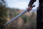 Steel arming sword with a lobed pommel, brown leather grip and straight guard held point down by a wrapped hand, wooded hills behind
