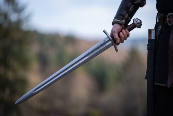Steel arming sword with a lobed pommel, brown leather grip and straight guard held point down by a wrapped hand, wooded hills behind