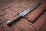 Steel arming sword with an olive green leather grip, blackened curved guard and faceted pommel laid on red brick paving, brown leather scabbard beside