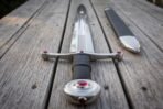 Steel arming sword with red stone set cross guard and wheel pommel, black leather grip, on grey decking with steel mounted scabbard