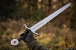 Steel arming sword with red stone set guard and wheel pommel held up by a gloved hand against autumn woodland