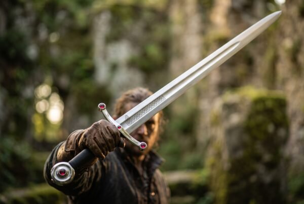 Steel arming sword with red set stones on the curved guard and disc pommel, etched knotwork band on the blade, held out in mossy woodland
