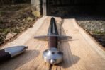 Steel arming sword seen from the pommel end along the fullered blade on an oak plank, brown leather scabbard with steel chape at the side