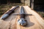 Close up of a steel arming sword wheel pommel and cross guard with a brown leather grip on an oak plank, scabbard chape beside