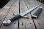 Close up of a steel arming sword wheel pommel set with a red stone, black leather grip and steel mounted scabbard on grey decking