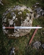 Steel arming sword with brown leather grip and engraved disc pommel lying on a mossy boulder beside its scabbard