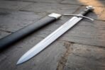 Steel arming sword with curved crossguard and wheel pommel lying beside its black leather scabbard on stone paving