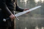 Steel arming sword with downswept crossguard and etched knotwork fuller held across both hands beside a lake