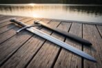 Steel arming sword with downswept guard lying beside a black leather scabbard on a wooden jetty at sunset