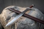 Steel arming sword with a fullered blade, brown leather grip and wheel pommel resting on a boulder across its brown scabbard