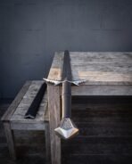 Steel barbed sword with faceted pommel laid across the corner of a weathered wooden table, spiked guard and black scabbard in view
