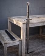 Steel barbed sword with spiked crossguard and faceted pommel standing point down beside a grey wooden table, black scabbard on the bench
