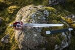 Steel basket hilt broadsword with red liner and black scabbard laid across a moss covered boulder in open hill country