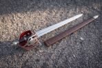 Steel basket hilt broadsword with red lined pierced guard and black wire bound grip on gravel beside a brown scabbard
