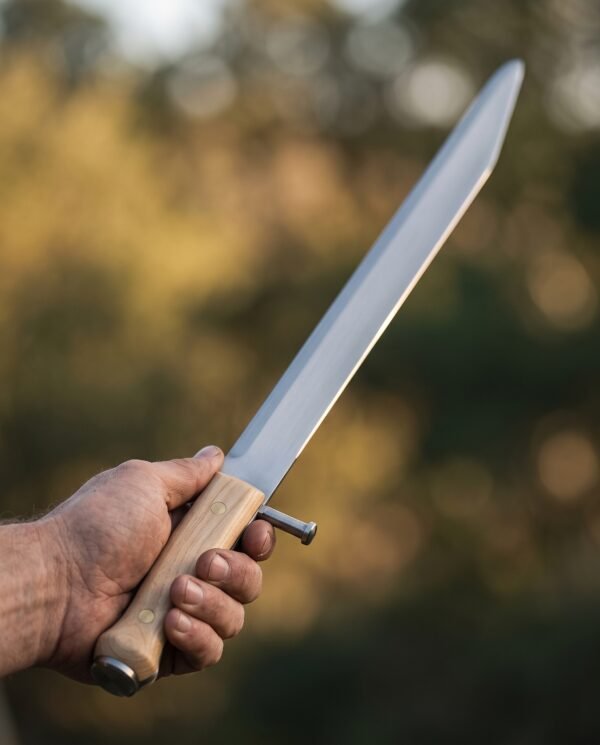 Steel bauernwehr knife with a pale wood scale grip and side nagel held in hand, clipped point blade against blurred autumn trees