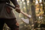 Steel bearded axe with an etched rune band and knotwork cheeks on a pale ash haft, carried at the hip by a man in a wool tunic in autumn woods