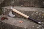 Steel bearded axe with etched rune band laid across mossy stone steps, pale ash haft, leather wrapped grip and a brown leather sheath behind