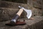 Steel bearded axe head with etched knotwork, pale ash haft with a dark leather grip wrap, leaning on stone steps beside its leather sheath