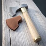 Steel bearded axe with a rune engraved cheek, brass collar and pale ash haft laid on grey weathered planks beside a tan leather blade sheath
