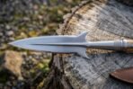 Steel boar spear head with swept lugs and engraved collar, close up on a weathered stump beside a leather sheath