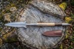 Steel boar spear with lugged head, pale wood and dark wood haft and butt spike laid on a stump next to its sheath