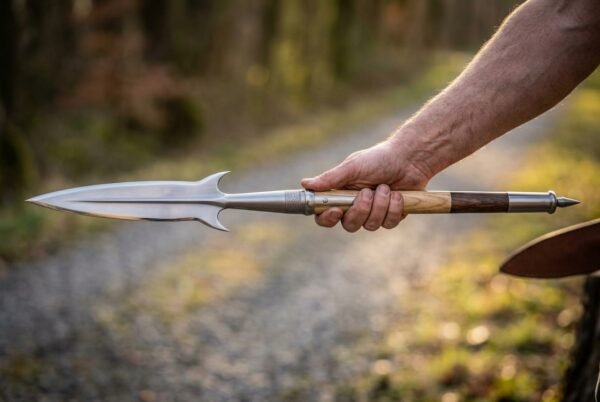 Steel boar spear with winged head and two tone wooden haft held out in one hand above a woodland path
