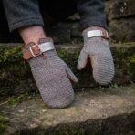 Steel chainmail mitten with tan leather cuff and knotwork buckle plate worn on a hand, the second mitten resting on a stone step behind