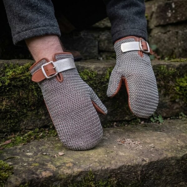 Steel chainmail mitten with tan leather cuff and knotwork buckle plate worn on a hand, the second mitten resting on a stone step behind
