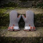Two steel chainmail mittens with tan leather trim and buckled cuffs standing side by side on a moss covered stone ledge