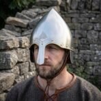 Steel conical nasal helmet with a pointed skull, brass rivets and a valknut engraving worn by a bearded man against a dry stone wall